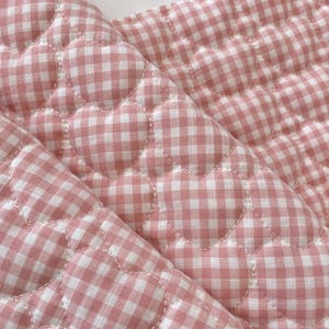 Könnte beinhalten: Nahaufnahme eines gesteppten Stoffes mit einem rosa-weißen Vichy-Muster. Der Stoff ist mit herzförmigen Steppungen versehen, wodurch eine strukturierte Oberfläche entsteht. Der Stoff ist gefaltet und zeigt das Muster und die Textur.