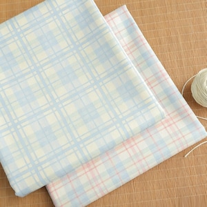 Puede incluir: Dos piezas de tela dobladas con un estampado de cuadros. Una es azul claro y blanca, la otra rosa y blanca. Una madeja de hilo blanco está en la esquina superior derecha. Las telas están sobre una superficie marrón claro.