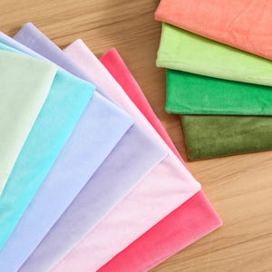 Puede incluir: Una colección de cuadrados de tela doblados en una variedad de colores sólidos que incluyen melocotón, verde claro, verde esmeralda, verde oliva, lavanda, azul claro, rosa pálido y rosa brillante, todos con una textura suave, afelpada y aterciopelada.