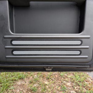 May include: A dark grey vehicle door panel featuring two horizontal, textured grey strips. The door has a recessed handle area. The background shows grass and leaves.