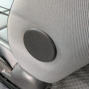 May include: A close-up of a black, circular, foam seat plug installed on a gray, vertically striped car seat. The seat has a worn appearance.