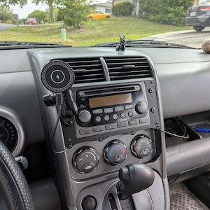 Op de afbeelding: Een auto dashboard met een draadloze oplaadpad. De pad is zwart en rond met een bliksemschicht symbool. Het dashboard heeft ook een radio, ventilatieopeningen en klimaatregelknoppen. Een klein decoratief beeldje staat op het dashboard.