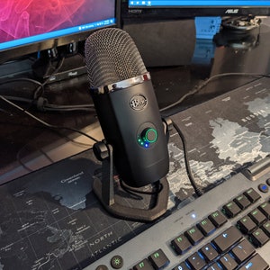 May include: A black Blue Yeti USB microphone with a green light on the knob. The microphone is on a black stand and is connected to a computer.