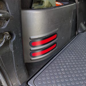 May include: A dark grey vehicle interior panel featuring two red stripes. The panel is part of a vehicle's interior, with a textured floor mat below. The panel has a curved design with the red stripes.
