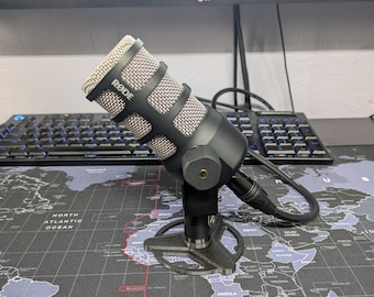 May include: A black RØDE microphone with a silver mesh grille, mounted on a black tripod stand. The microphone is connected to a black cable.
