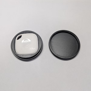 May include: A black 3D printed case for a Tile Mate Bluetooth tracker. The case has a textured surface and a raised lip around the edge. The Tile Mate is white with a black logo and a small hole for a keyring.