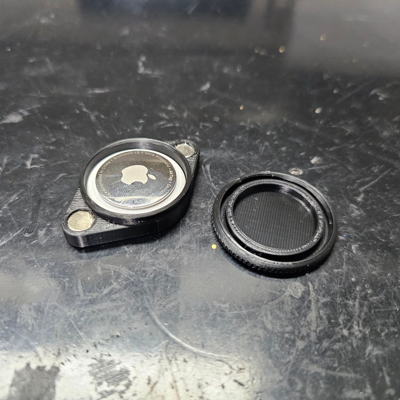 May include: A black plastic case with a silver Apple AirTag inside. The case has a small magnet on the side.