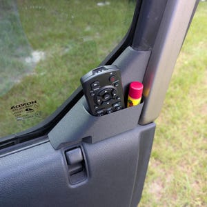 May include: A black plastic car door storage pocket containing a black remote control with multiple buttons and a small cylindrical lip balm tube with a bright pink cap, positioned against the interior panel of a vehicle door.