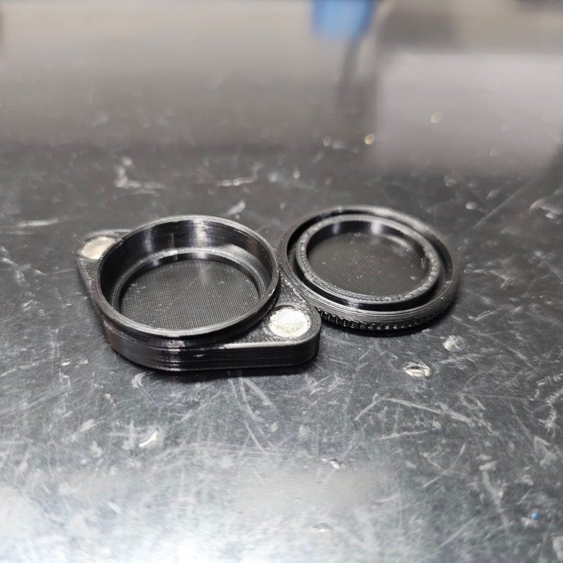 May include: A black plastic container with a lid. The container has a round opening and a textured surface. The lid has a smooth surface and a slightly raised edge.
