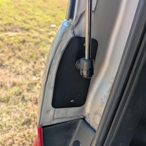 May include: Close-up of a car trunk interior. A black, rectangular panel is mounted on the silver metal frame. A black gas strut is attached to the panel and the frame. The background shows green grass.