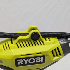 Peut inclure: Gros plan sur un outil électrique Ryobi jaune et noir. L'outil est équipé d'un tuyau noir et d'un bouton noir. Le logo Ryobi est visible sur le côté. L'outil présente un design nervuré et un accent gris.