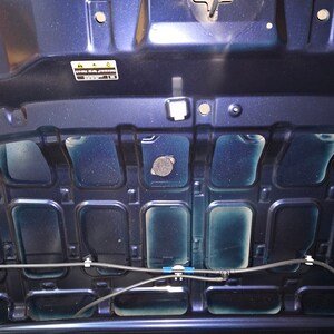May include: The underside of a dark blue car bonnet with a black plastic cover and a warning label that reads "Caution: Hot Surface".