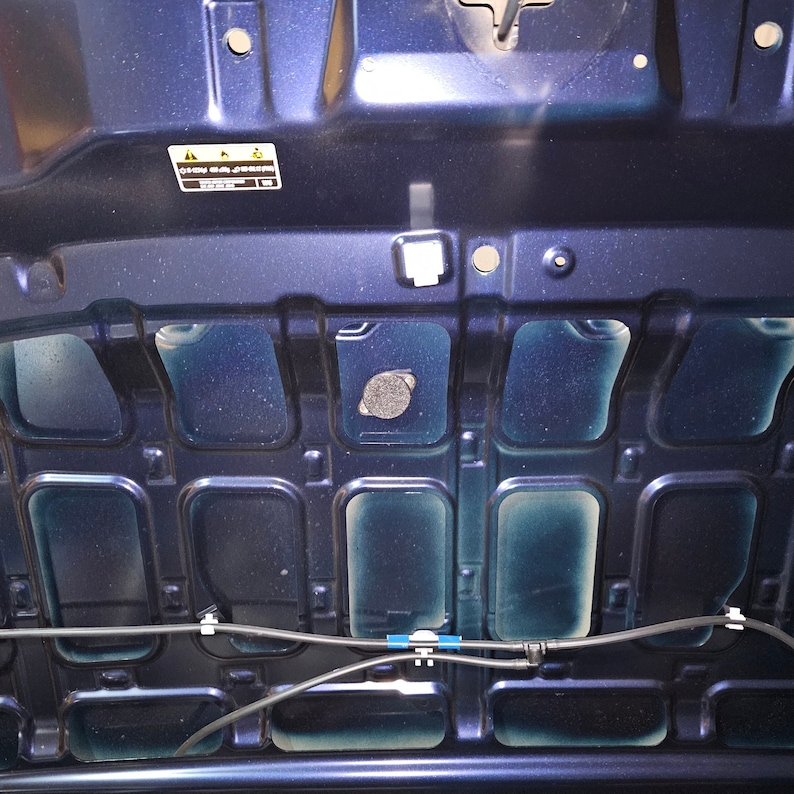 May include: The underside of a dark blue car bonnet with a black plastic cover and a warning label that reads "Caution: Hot Surface".