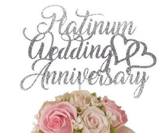 Może przedstawiać: Biały tort ozdobiony różowymi różami i srebrnym, błyszczącym topperem na tort z napisem "Platinum Wedding Anniversary".