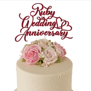 May include: A round, cream-coloured cake topped with pink and white roses and a red glitter topper reading "Ruby Wedding Anniversary." The cake sits on a light pink base with a ribbon and pearl trim.
