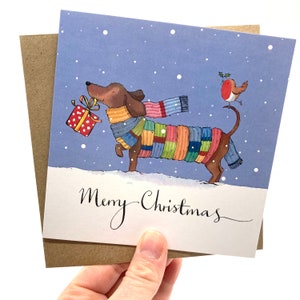 May include: A Christmas card featuring a brown dachshund wearing a colorful scarf and carrying a red and white polka dot gift box. A red robin with a holly sprig is perched on the dog's tail. The card has a blue background with white snowflakes and the text "Merry Christmas" in black cursive.