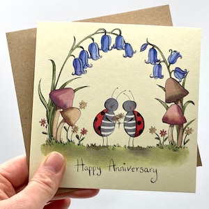 May include: A greeting card with a hand-painted illustration of two ladybugs holding a bouquet of flowers. The ladybugs are standing under an arch of bluebells and mushrooms. The card reads "Happy Anniversary".