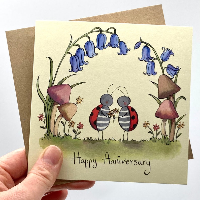 May include: A greeting card with a hand-painted illustration of two ladybugs holding a bouquet of flowers. The ladybugs are standing under an arch of bluebells and mushrooms. The card reads "Happy Anniversary".