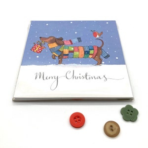 May include: A Christmas card featuring a cartoon dachshund wearing a colorful scarf and carrying a wrapped gift. A robin perches on a branch above the dog. The card has a blue background with white snowflakes and the text "Merry Christmas" in cursive.