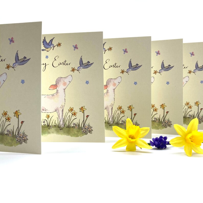 May include: Five blank greeting cards with a pastel yellow background. Each card features a white lamb with a blue bird flying above it. The cards have the text "Happy Easter" printed on them. The cards are arranged in a row with the text facing the viewer. There are yellow daffodils and blue flowers in the foreground.