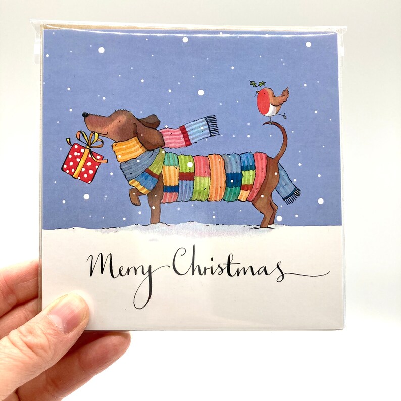 May include: A Christmas card featuring a cartoon dachshund wearing a colorful scarf and carrying a red and yellow gift box. A red robin perches on the dog's tail. The card has a blue background with white snowflakes and the text "Merry Christmas" in black cursive.
