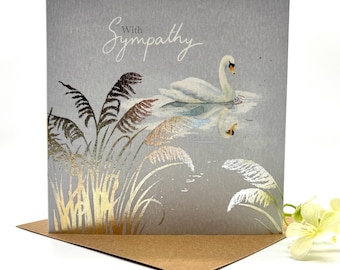 May include: A square greeting card with a gray background featuring a white swan swimming on water, reflected below. The card displays the text With Sympathy in white script. Gold foil accents depict tall reeds and grasses in the foreground.