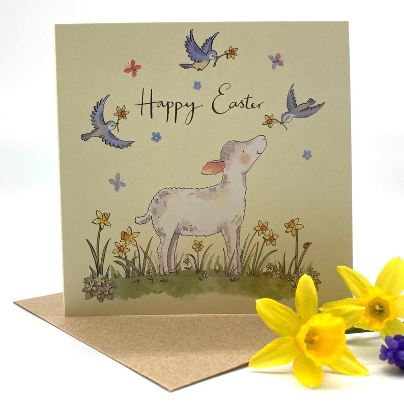 May include: A light yellow card with a white lamb standing in a field of yellow daffodils. Two blue birds with pink accents are flying above the lamb. The card says "Happy Easter" in cursive.