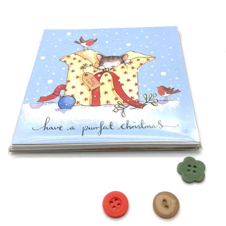 May include: A Christmas card with a blue background and a white cat peeking out of a yellow and red gift box with red stars. The cat is holding a red ribbon. Two red robins are perched on the gift box and on a branch. The card reads "have a purrfect christmas".