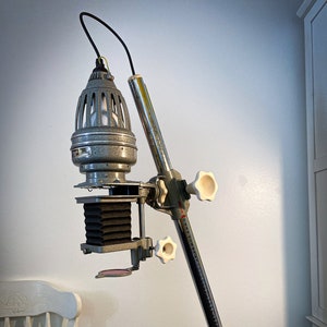 May include: A vintage silver and black enlarger with a white lampshade and a green base. The enlarger is mounted on a silver metal arm with a white knob.
