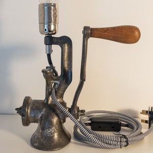 May include: A vintage-style metal meat grinder lamp with a wooden handle, black and white patterned cord, and a black plug. The lamp has a silver light socket and a clear light bulb.