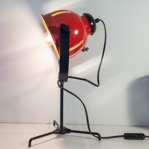 May include: A red and black adjustable desk lamp with a tripod base and a round shade. The lamp is turned on and the light is shining.