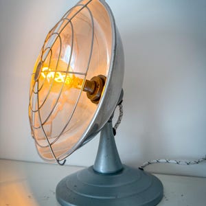 May include: A table lamp with a circular, metallic, bowl-shaped reflector and a protective wire cage. The lamp features a central, glowing amber-colored filament bulb and a solid, circular base in a muted blue-gray finish, connected by a cord.