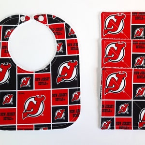 May include: A set of four red and black fabric burp cloths and a bib featuring the New Jersey Devils logo. The fabric has a repeating pattern of the logo and the words "New Jersey Devils".