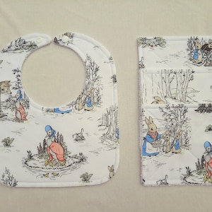 May include: A white baby bib and matching burp cloth set. Both items feature a black and white illustrated scene with rabbits, ducks, and other animals. The bib has a rounded shape with snap closures. The burp cloth is rectangular and quilted.