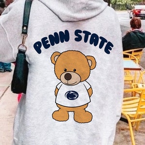 Könnte beinhalten: Ein grauer Hoodie mit einem Comic-Bären, der ein blau-weißes Shirt mit dem Text "Penn State" auf dem Rücken trägt.