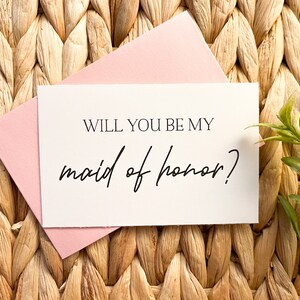 May include: A white card with black text that reads "Will you be my maid of honor?" The card is sitting on a woven wicker surface.