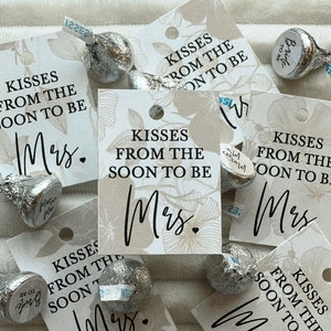 K&ouml;nnte beinhalten: Eine Sammlung kleiner wei&szlig;er Geschenkanh&auml;nger mit Blumendesigns und dem Text "Kisses from the soon to be Mrs." in Schwarz gedruckt. Die Anh&auml;nger sind auf einer wei&szlig;en Oberfl&auml;che mit silbernen, in Folie verpackten Schokoladenk&uuml;ssen verstreut.