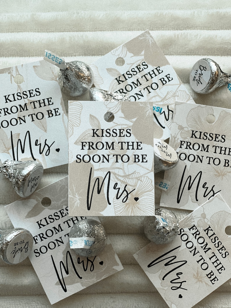 K&ouml;nnte beinhalten: Eine Sammlung kleiner wei&szlig;er Geschenkanh&auml;nger mit Blumendesigns und dem Text "Kisses from the soon to be Mrs." in Schwarz gedruckt. Die Anh&auml;nger sind auf einer wei&szlig;en Oberfl&auml;che mit silbernen, in Folie verpackten Schokoladenk&uuml;ssen verstreut.