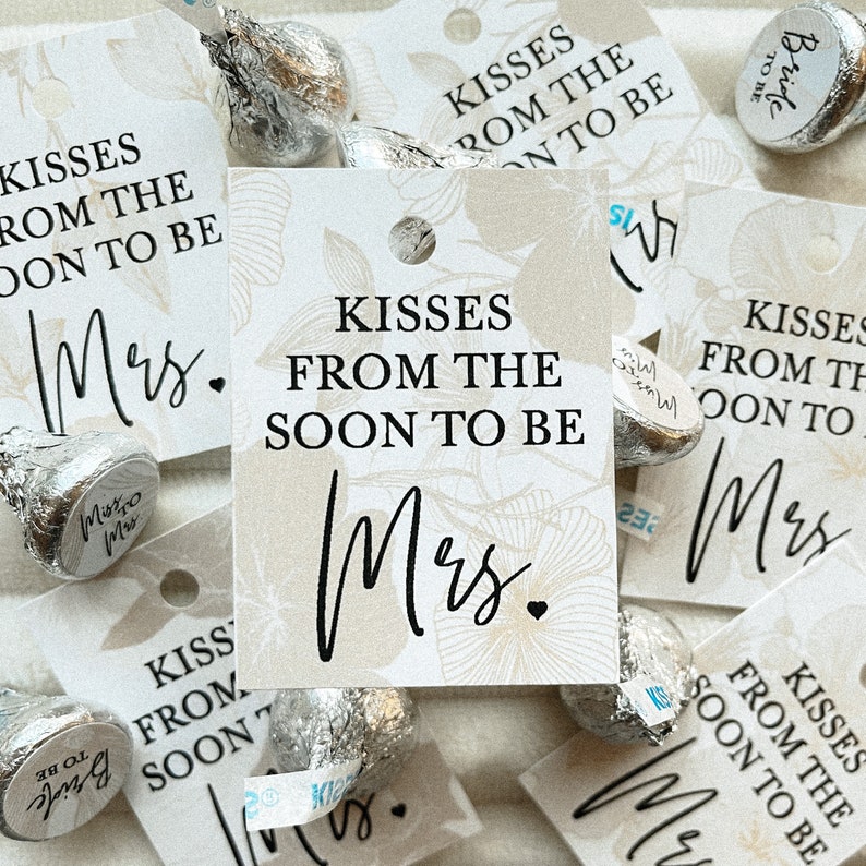 K&ouml;nnte beinhalten: Eine Sammlung kleiner wei&szlig;er Geschenkanh&auml;nger mit dem Text "Kisses from the soon to be Mrs." in schwarzer Schrift. Die Anh&auml;nger sind mit einem floralen Muster verziert und an kleine Schokoladenk&uuml;sschen befestigt.