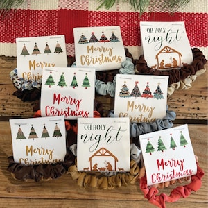 May include: Nine Christmas-themed scrunchies with different designs. The scrunchies are made of brown, blue, gold, and red fabric. The designs include Christmas trees, the words "Merry Christmas", and the words "Oh Holy Night".