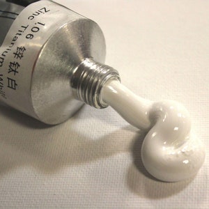 May include: A tube of white paint with the label "106 Titanium White" and "Zinc" in English and Japanese. The paint is being squeezed out of the tube, forming a white blob.