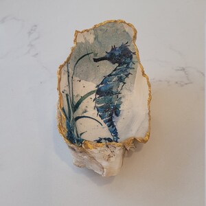 以下が含まれることがあります： 金色の縁取りが施された青と白で描かれた貝殻で、中には青いタツノオトシゴのイラストが描かれています。