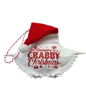 May include: A white seashell ornament with a red Santa hat. The ornament has a red and white string for hanging and says "I'm Dreaming of a Crabby Christmas" with red crabs and snowflakes.