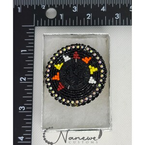 May include: A black beaded round pin with a paw print in the center. The pin is surrounded by a colorful beaded design with red, orange, yellow, and white beads. The pin is in a small white box with the text "Namewe Customs" on the bottom.
