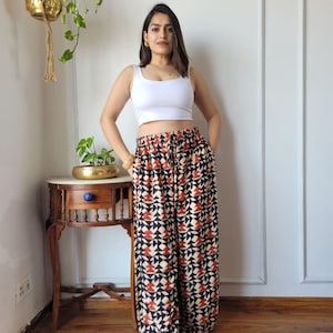 Op de afbeelding: Een vrouw draagt een witte tanktop en een palazzo broek met patroon. De broek heeft een geometrisch ontwerp in zwart, rood en crème. De outfit is casual en stijlvol, geschikt voor dagelijks gebruik.