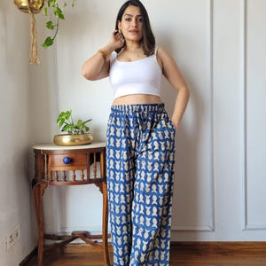 Peut inclure: Une femme porte un crop top blanc et un pantalon palazzo bleu. Le pantalon présente un motif répétitif de formes beige clair sur un fond bleu foncé. Elle se tient devant un mur blanc, avec une petite table en bois et des plantes suspendues.