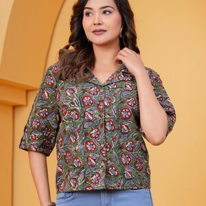 Könnte beinhalten: Ein kurzärmeliges, olivgrünes Hemd mit durchgehender Knopfleiste und einem sich wiederholenden Blumenmuster in Rot, Weiß und Blau, kombiniert mit einer hellblauen Jeans. Das Hemd hat einen klassischen Kragen und einen geraden Saum.