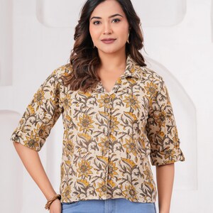 Puede incluir: Una camisa abotonada con base beige y un patrón floral repetitivo en tonos amarillo mostaza y gris oscuro. La camisa tiene un cuello clásico, mangas de tres cuartos con puños doblados y un dobladillo recto sobre unos pantalones vaqueros azules.
