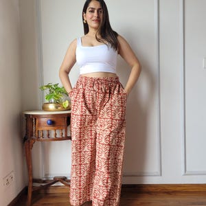 May include: A white sleeveless crop top paired with wide-leg trousers featuring a repeating rust-coloured botanical print on a cream background. The trousers have a high-waisted fit with a drawstring waistband and side pockets.