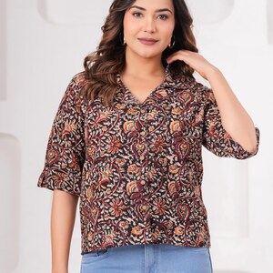 Puede incluir: Una camisa abotonada con base negra y un patrón floral y de cachemira denso y complejo en tonos naranja, crema y rojo oscuro. La camisa tiene cuello con solapas, mangas tres cuartos con puños doblados y pantalones vaqueros azul claro.